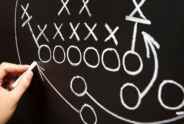 Hand diagramming football play in chalk on chalkboard.